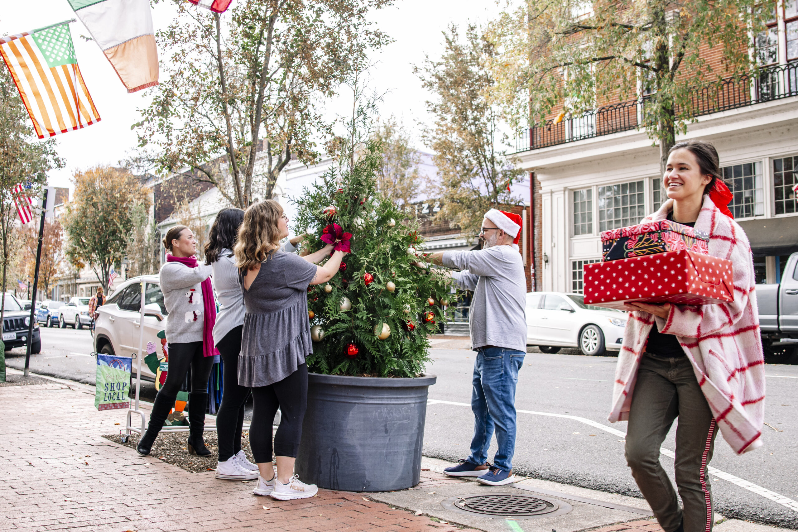 Image of people decorating a Christmas tree and a person walking by with a stack of presents. 